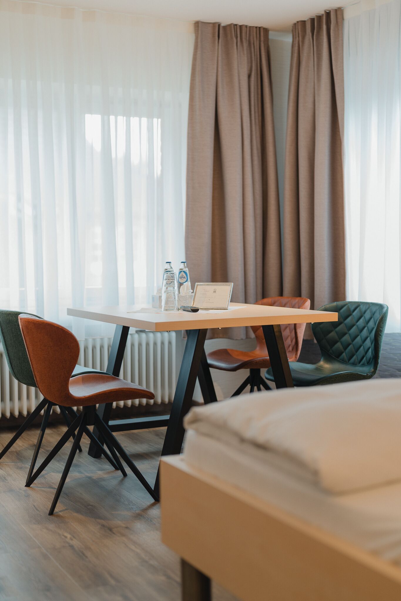Habitación de hotel con una cama, un escritorio con una computadora portátil, dos sillas y una ventana con cortinas.