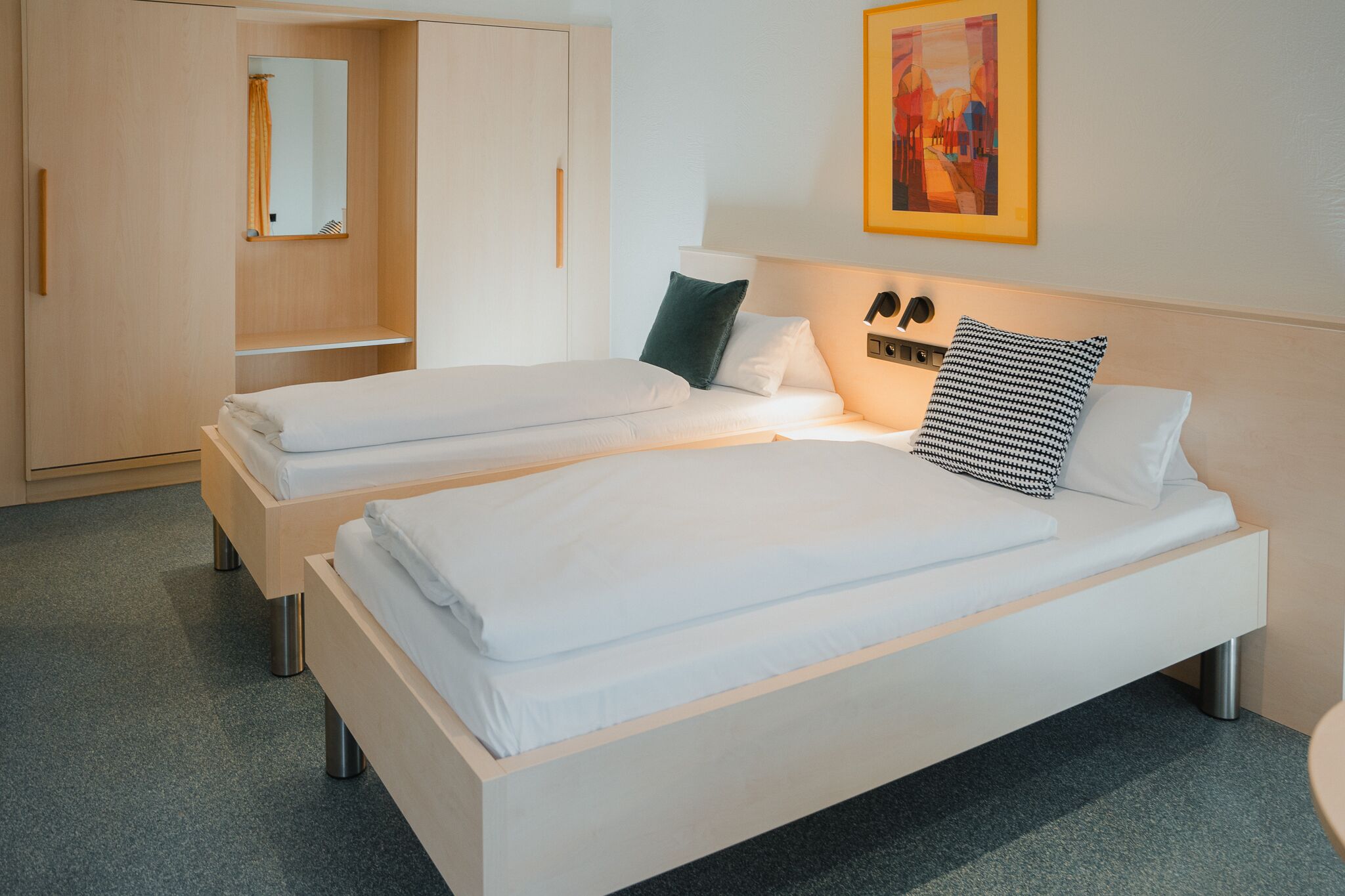 A hotel room with two single beds, a wardrobe, a mirror, and a painting on the wall.