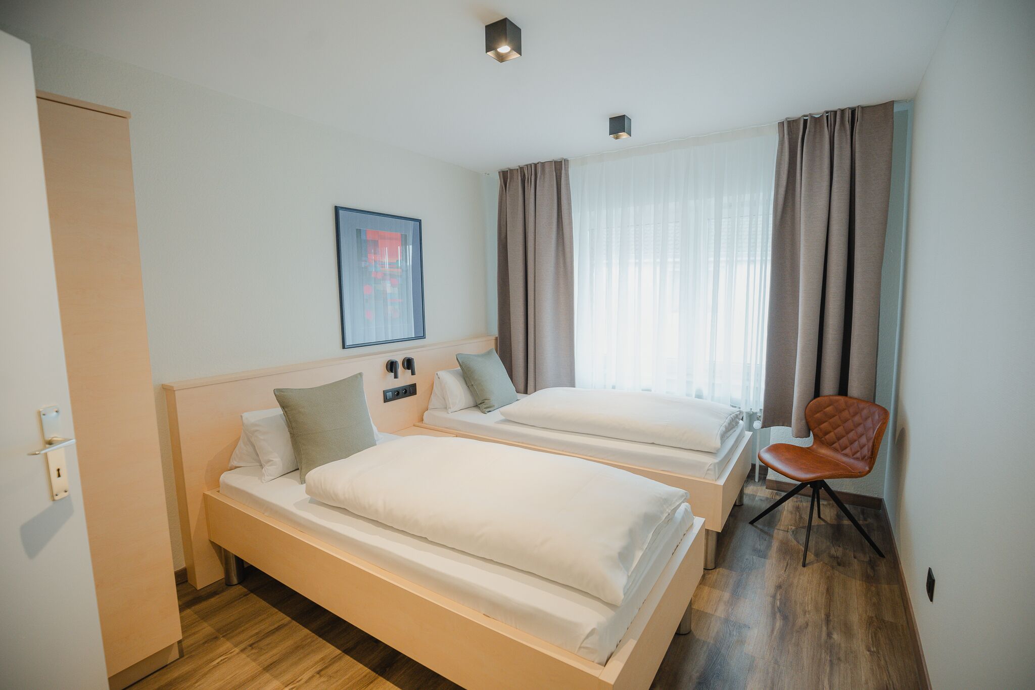 A hotel room with two beds, a chair, a painting, and a window with curtains.