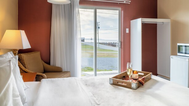 Ein Hotelzimmer mit Bett, Sessel, Tisch mit Speisen und Getränken, Lampe und Blick ins Freie durch eine Schiebetür.