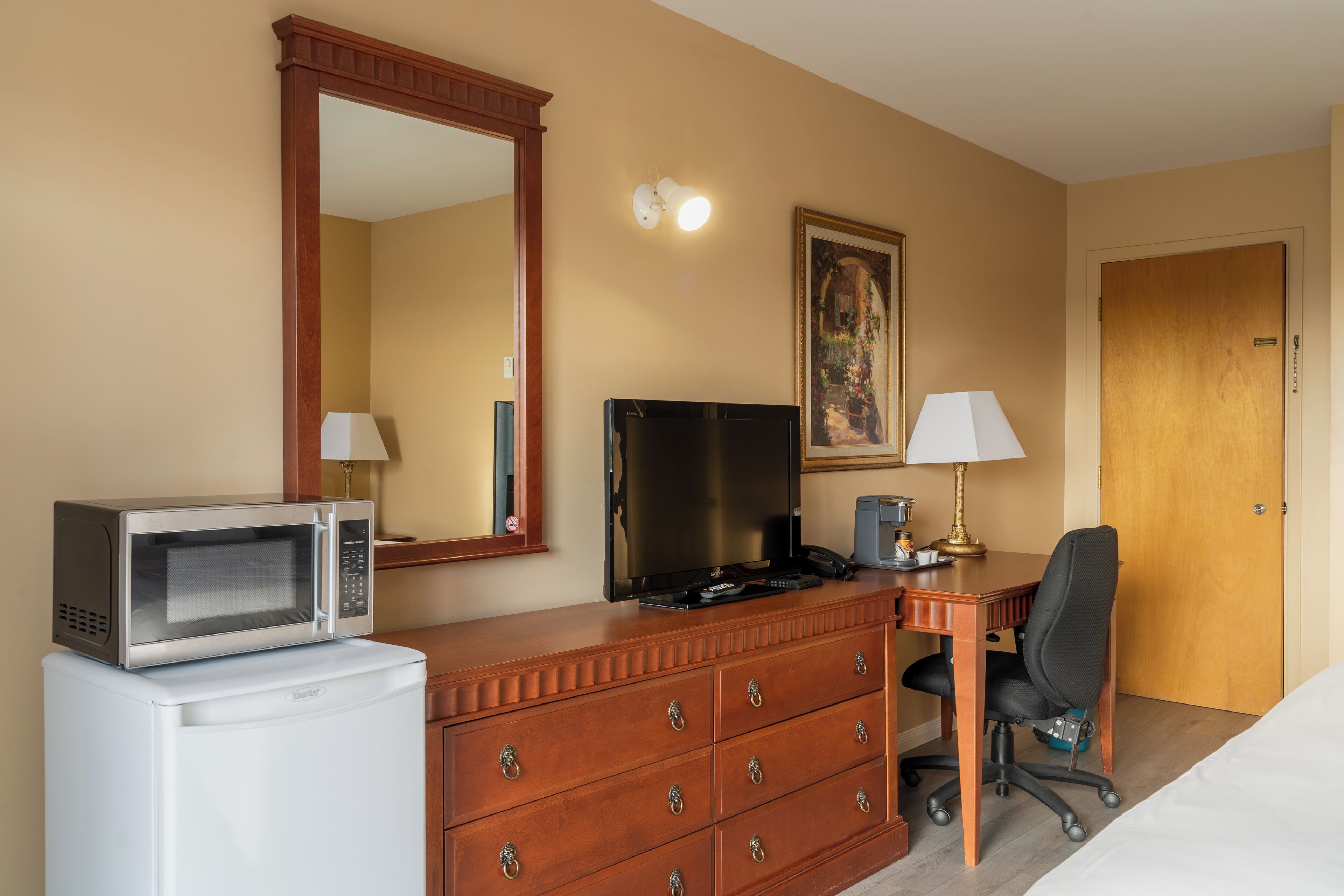A hotel room with a wooden dresser, a microwave, a television, a desk with a chair, a lamp, and a door.