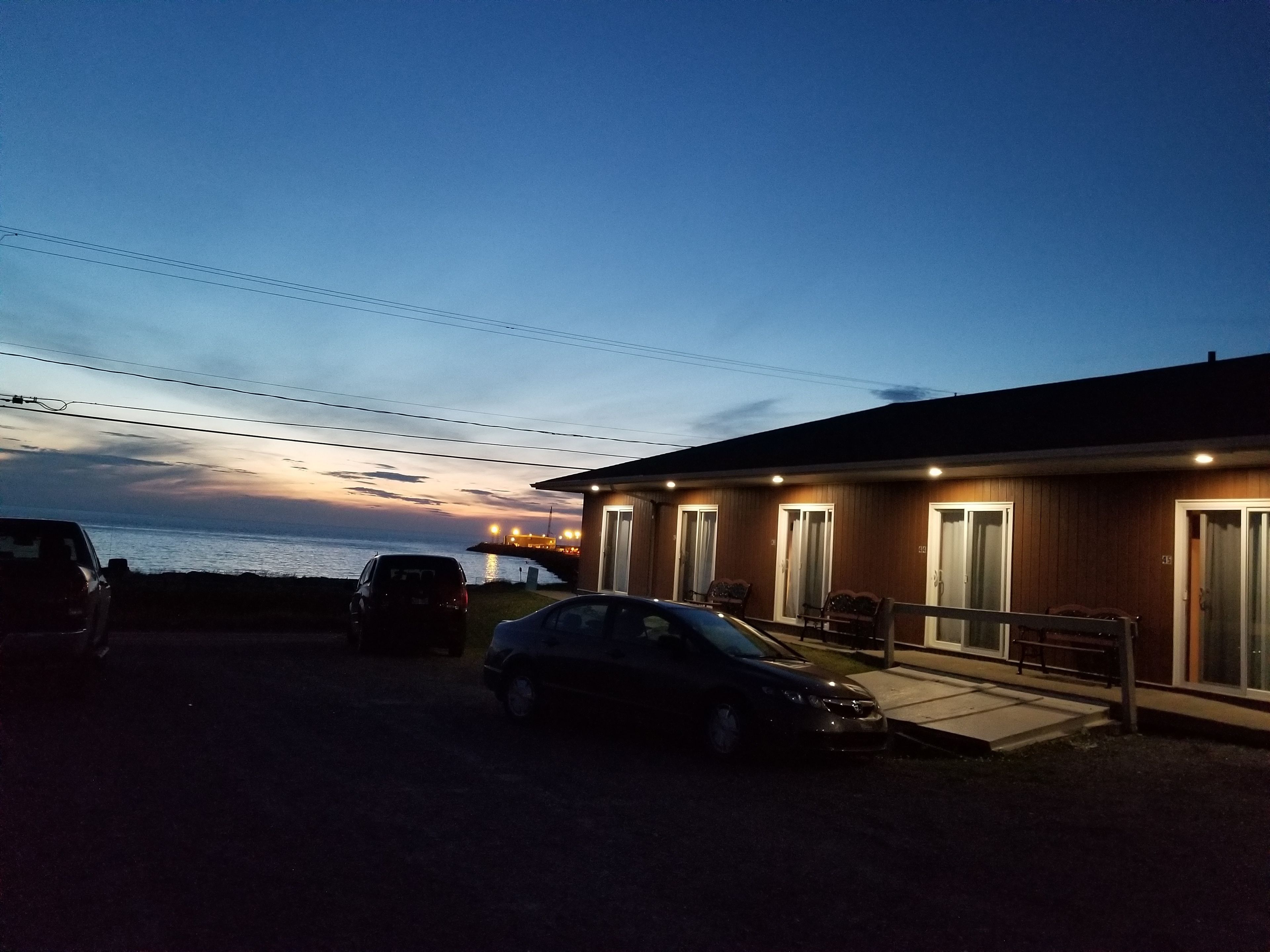 A building with a wooden exterior and multiple windows, a parking area with cars, and a view of the sea at dusk.