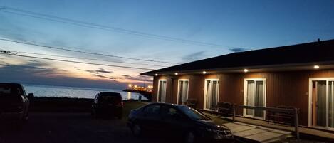 A building with a wooden exterior and multiple windows, a parking area with cars, and a view of the sea at dusk.