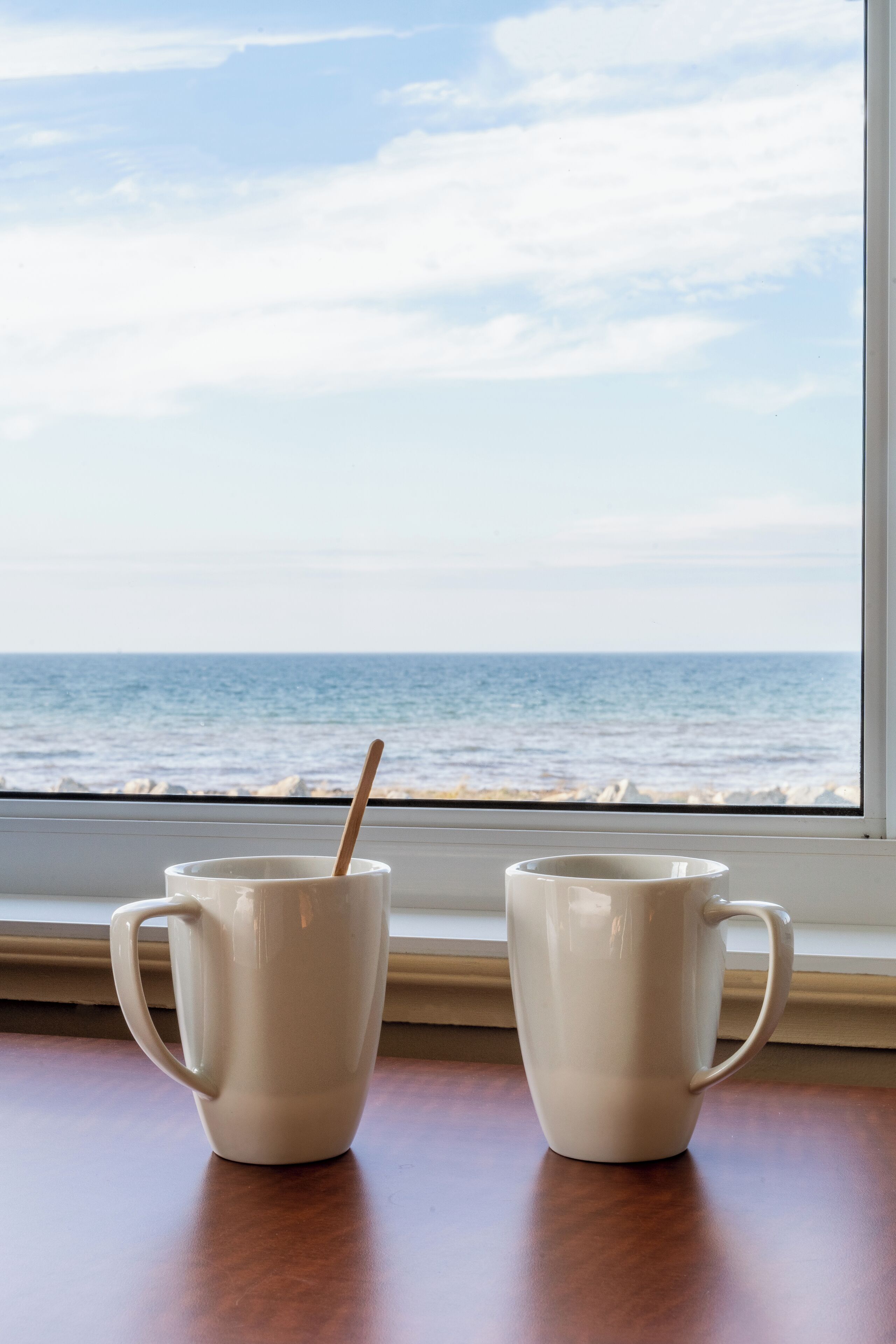 Twee witte mokken op een houten tafel bij een raam met uitzicht op de oceaan.