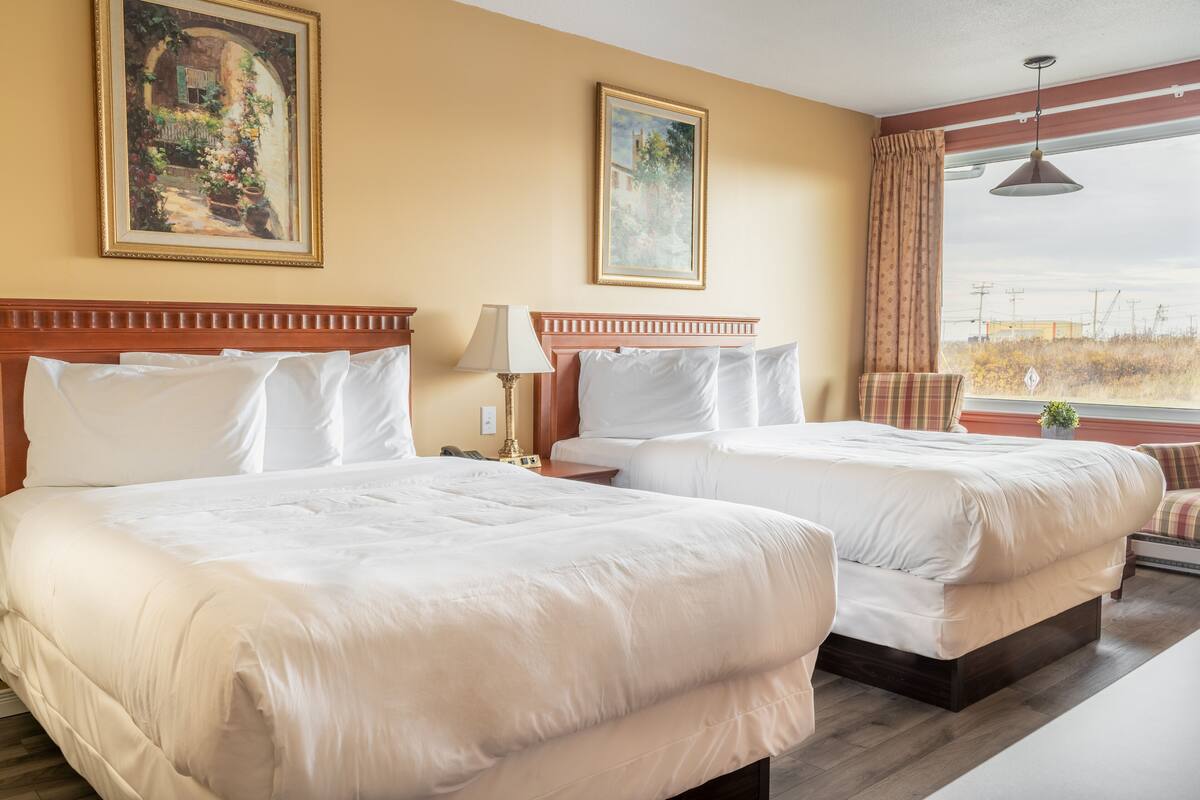 Quarto de hotel com duas camas, uma janela grande com cortinas e duas pinturas nas paredes.