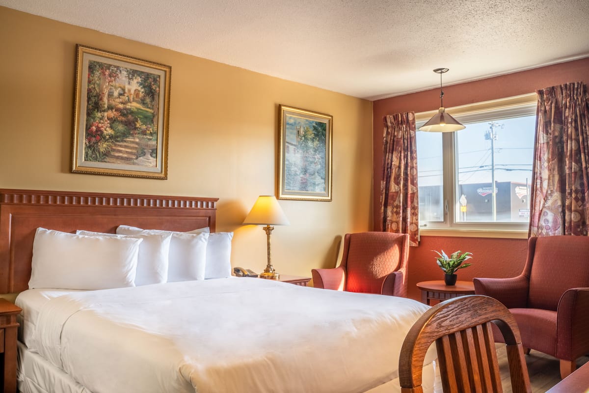 Quarto de hotel com uma cama grande, cabeceira de madeira, uma escrivaninha com abajur, uma cadeira e dois quadros na parede.