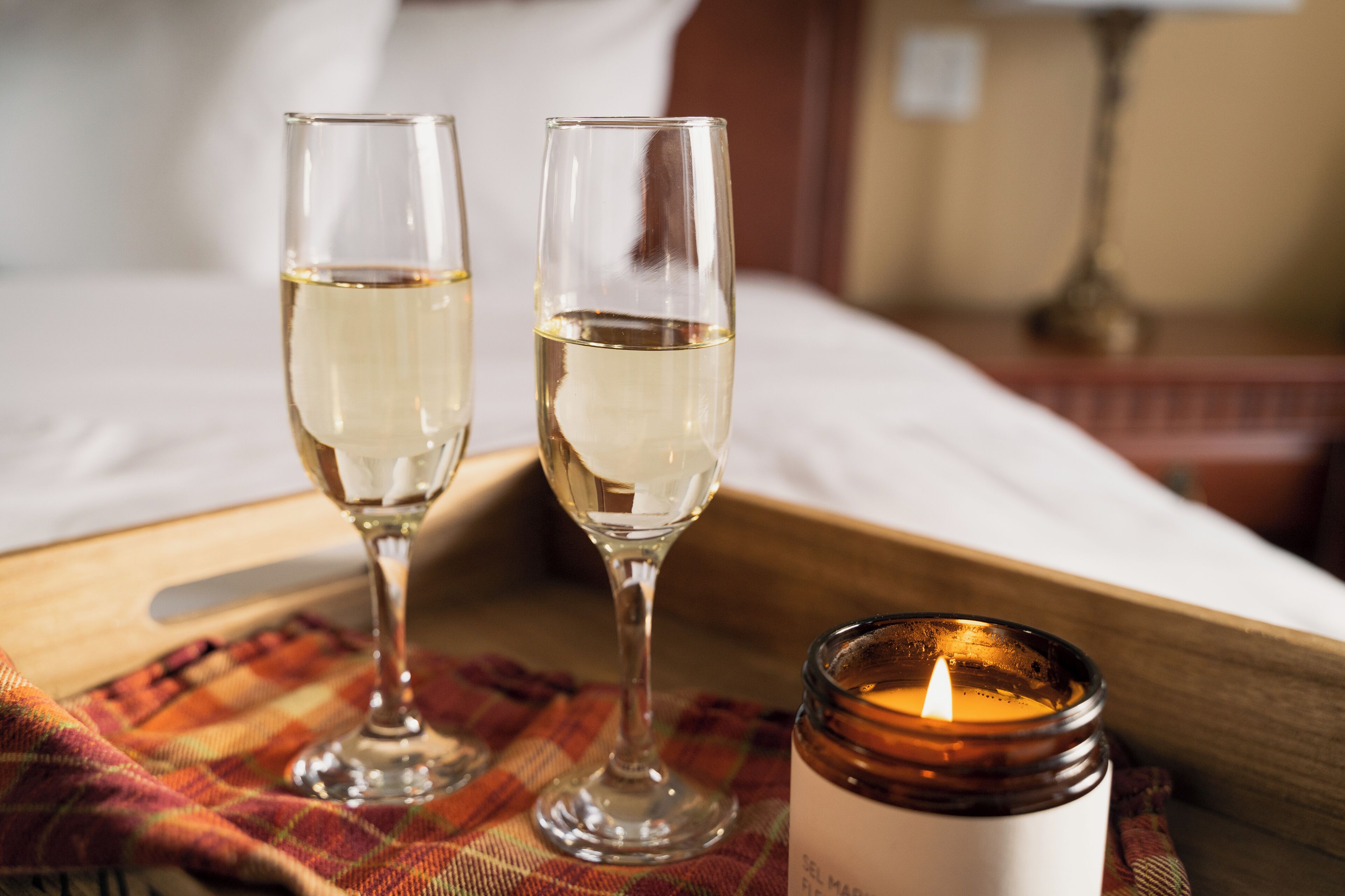 Two glasses of white wine on a tray, a lit candle, and a bed in the background.