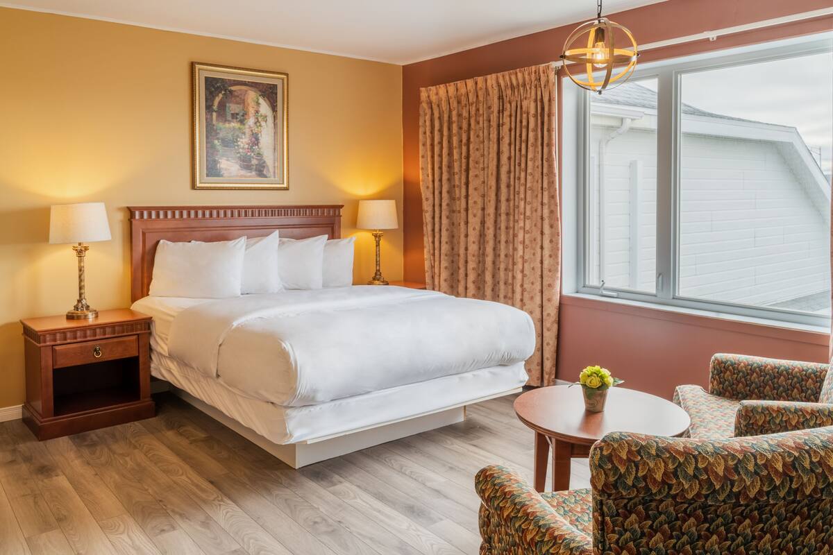 Quarto de hotel com uma cama grande, duas mesas de cabeceira com abajures, uma pequena mesa redonda com um vaso de flores, poltronas estampadas e uma janela com cortinas.
