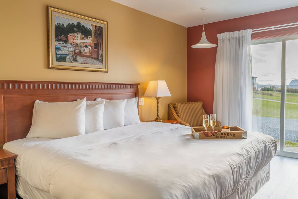 Quarto de hotel com cama grande, quadro na parede, luminária e bandeja com copos e garrafa.