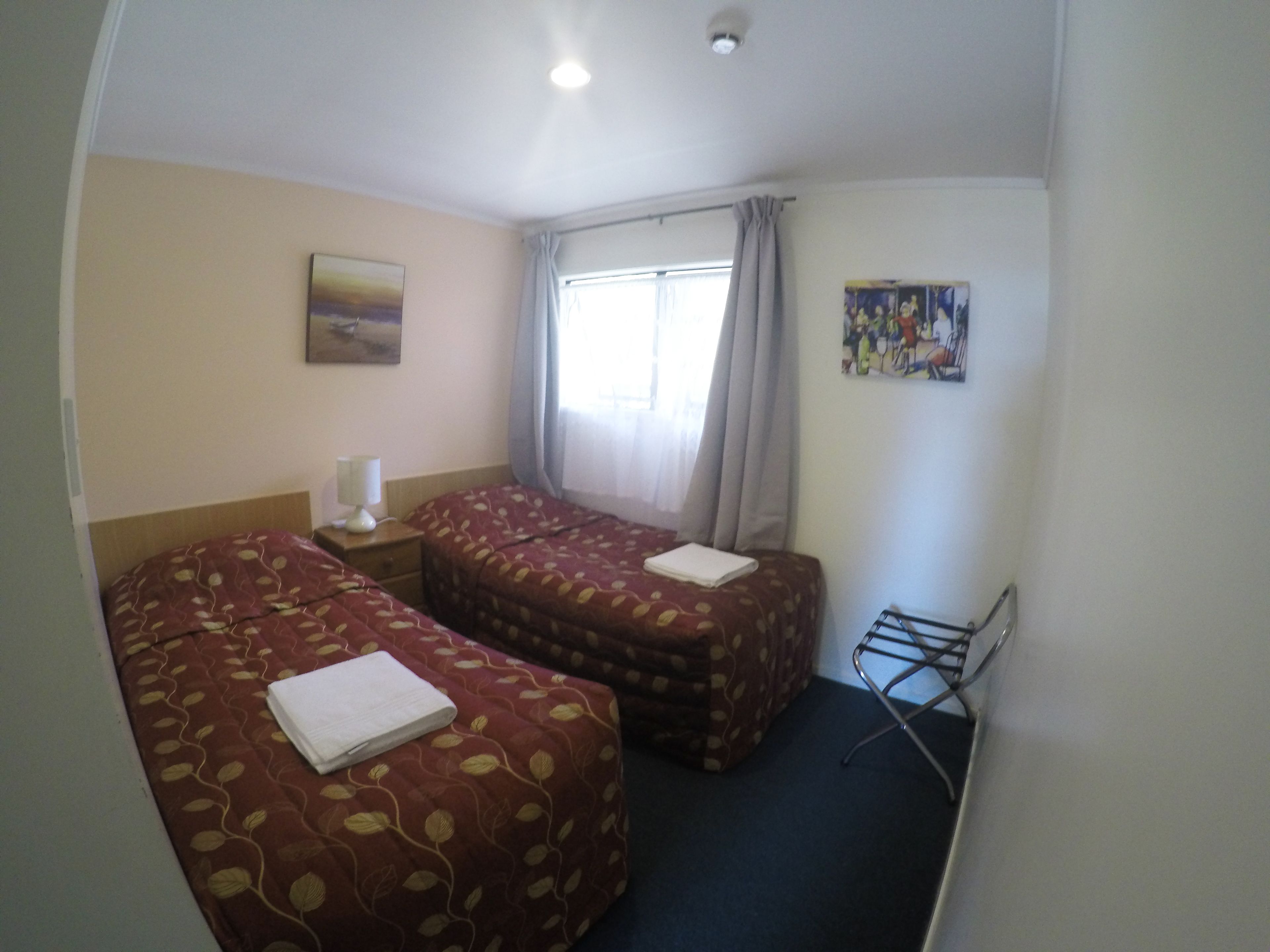 Quarto de hotel com duas camas, mesa de cabeceira, janela com cortinas, um quadro na parede e um porta-toalhas.
