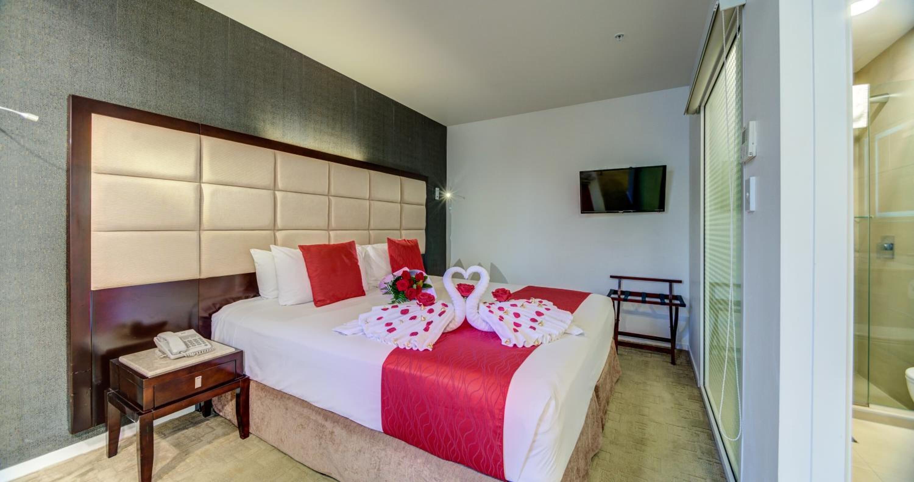 A hotel room with a large bed, a bedside table with a phone, a TV mounted on the wall, and a shower area visible through a glass door.