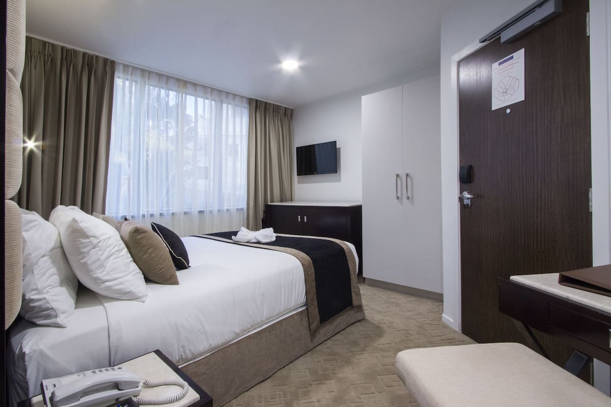 A hotel room with a bed, bedside table, TV, wardrobe, and a desk with a chair.