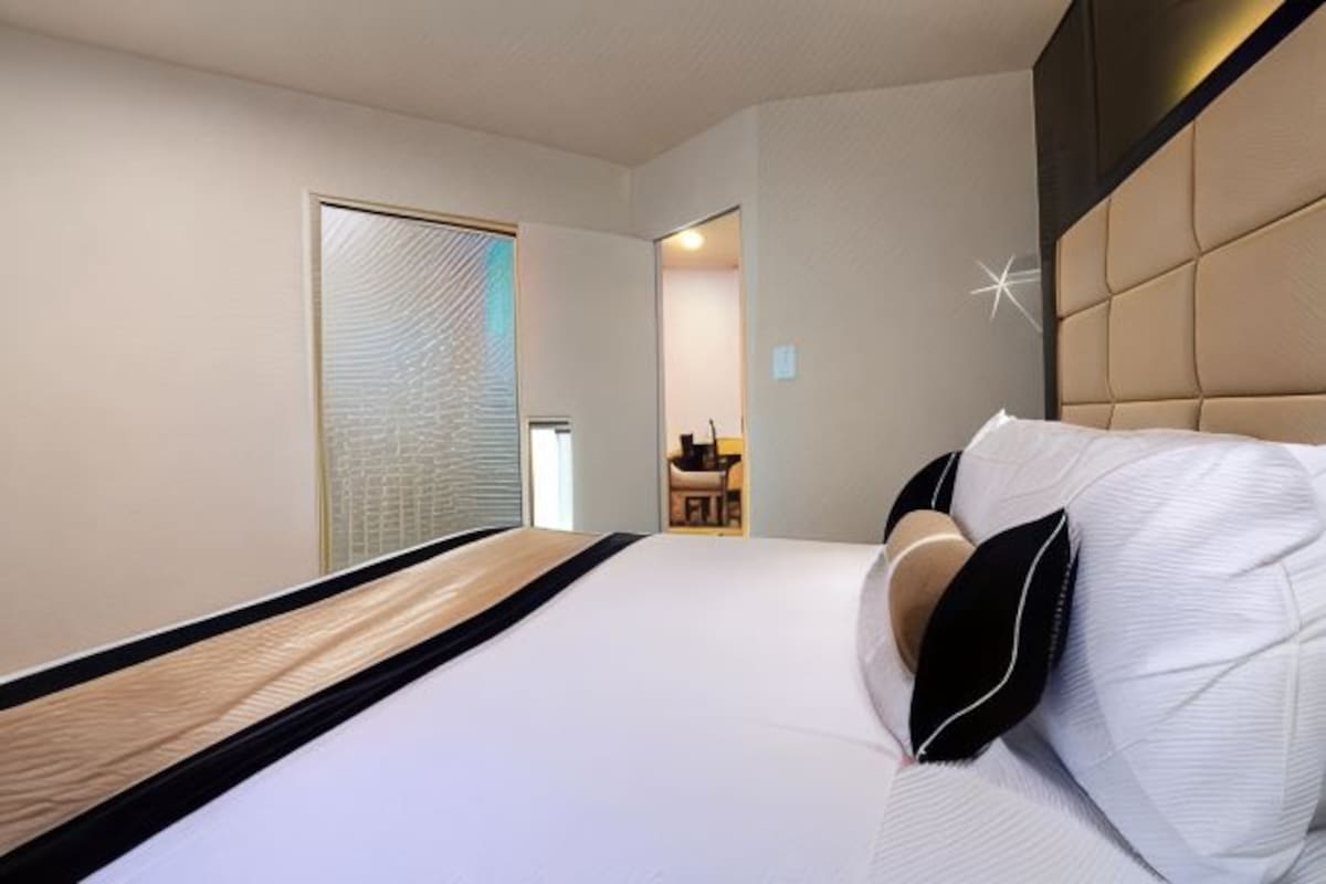 A hotel room with a bed, a headboard, a door, and a small table with a chair.