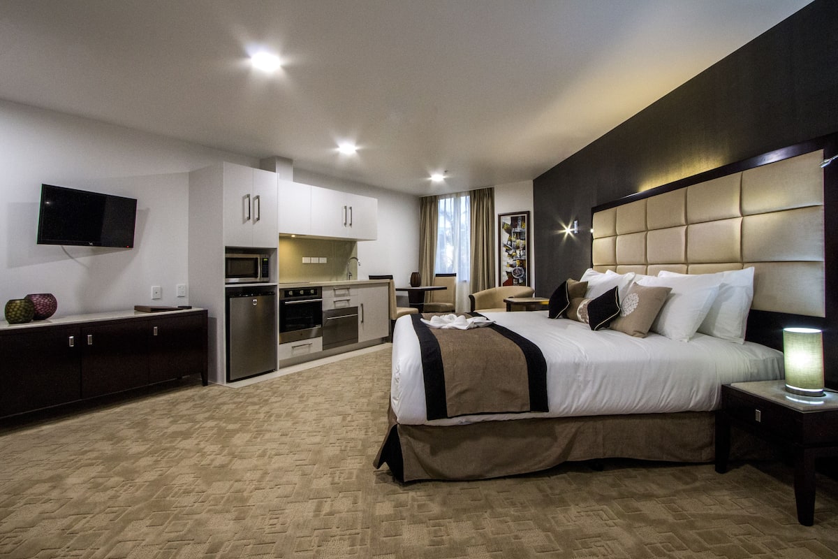 A modern hotel room with a large bed, a flat-screen TV, a kitchenette, and a bathroom.