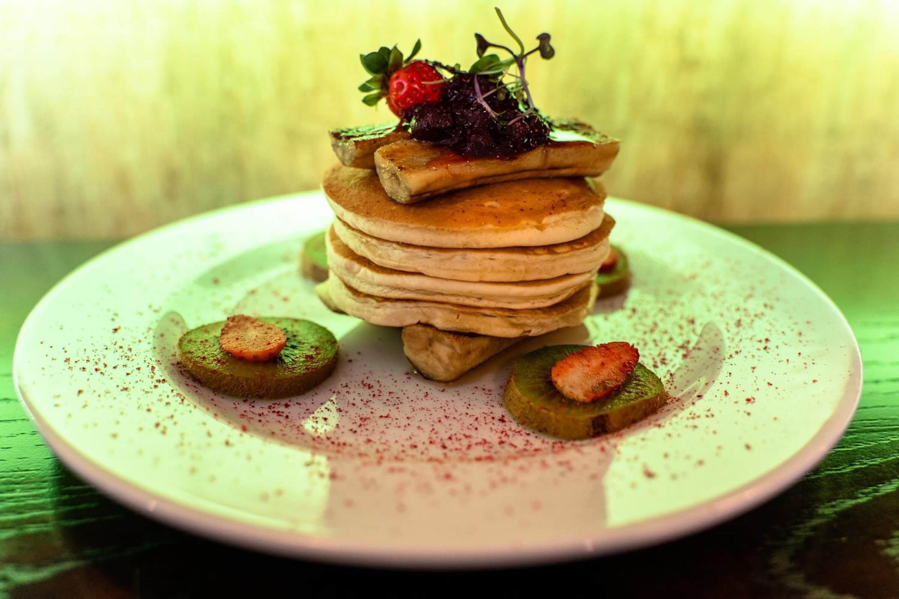 A stack of pancakes with a strawberry and a slice of kiwi on a plate.