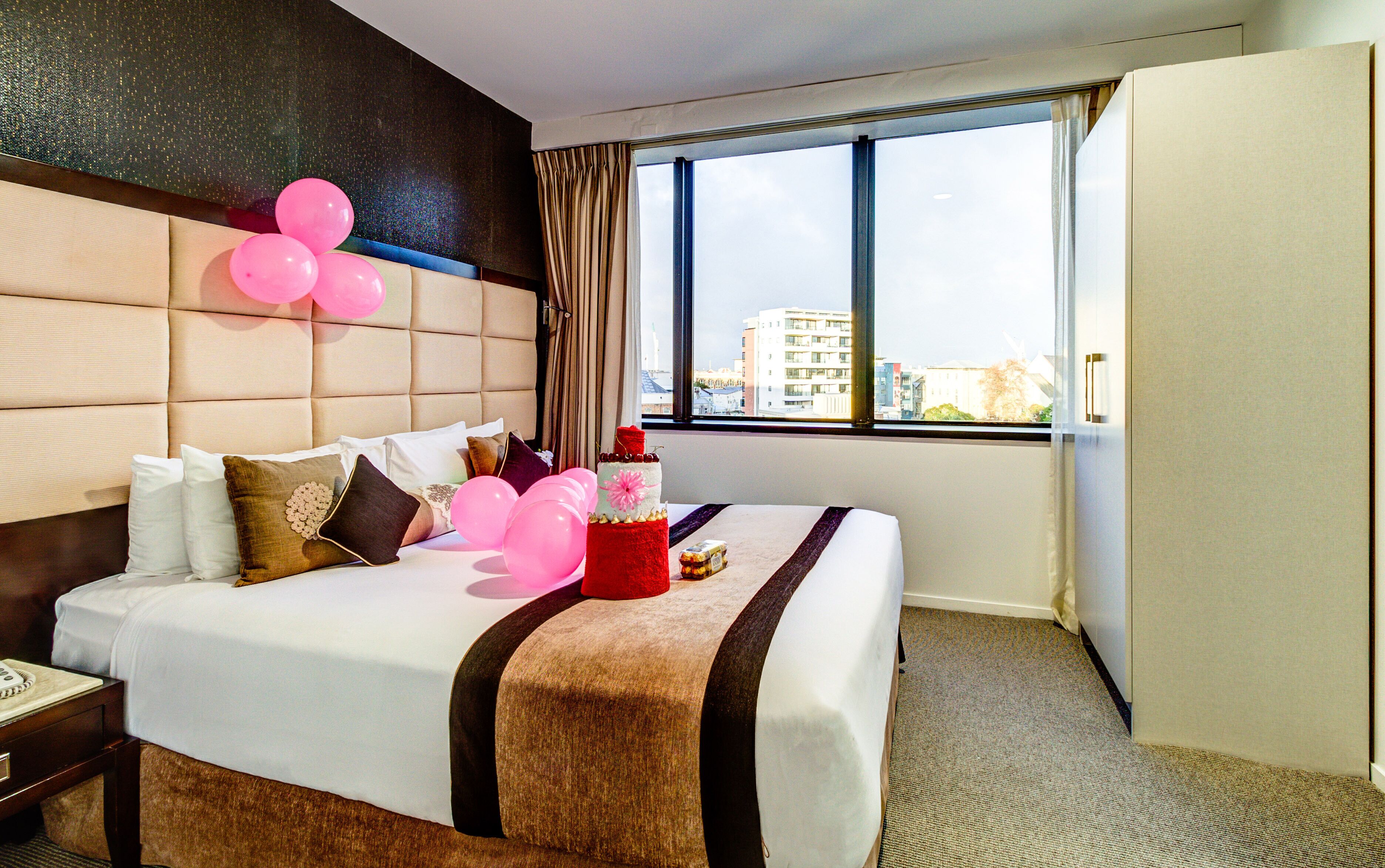 A hotel room with a large bed, a headboard, a window with curtains, and a view of buildings outside.