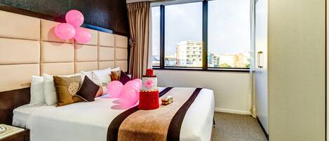 A hotel room with a large bed, a headboard, a window with curtains, and a view of buildings outside.
