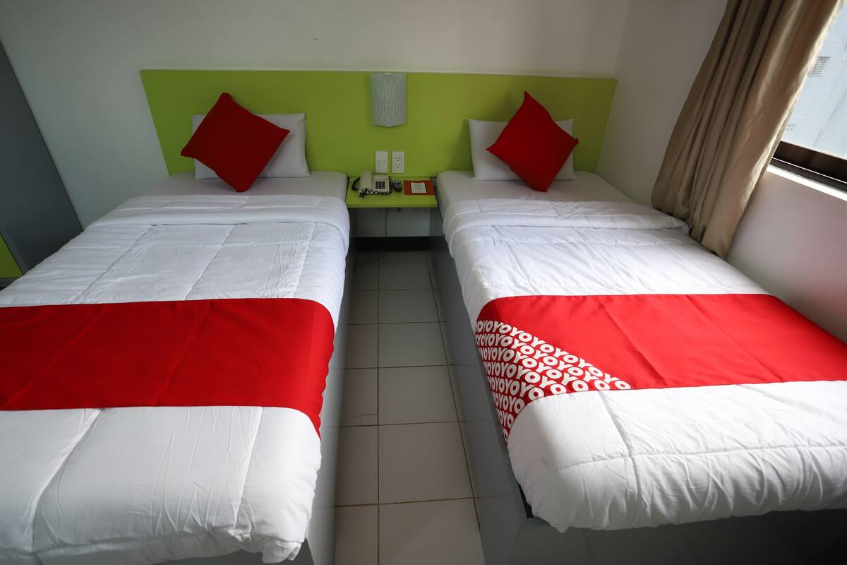 A hotel room with two beds, each with white and red bedding, a nightstand with a phone and a lamp, and a window with curtains.