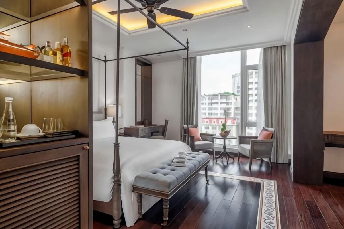 A hotel room with a four-poster bed, a seating area with a table and chairs, and a view of the cityscape through large windows.