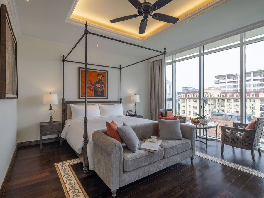 Amplia habitación de hotel con cama con dosel, sofá gris, comedor con mesa y sillas, y ventanas grandes con vistas a la ciudad.