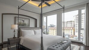 Un dormitorio amplio con una cama grande, un banco, un ventilador de techo y vistas a la ciudad a través de amplias ventanas.