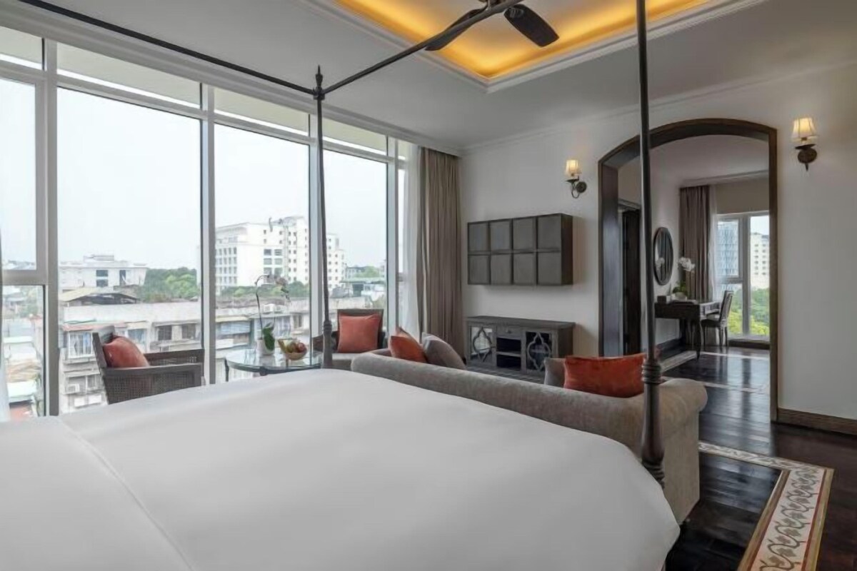 A modern hotel room with a large bed, a sofa, a small table, and a view of the city through large windows.