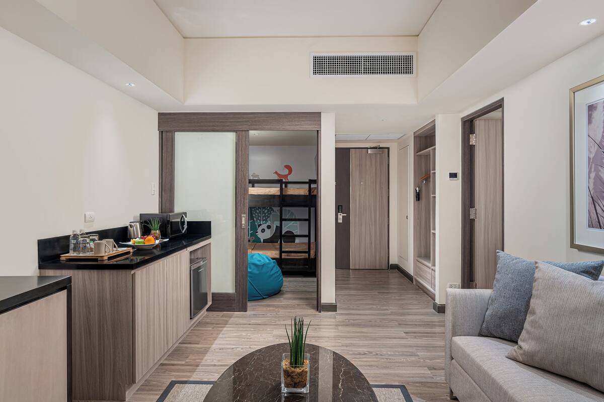 A modern apartment with a kitchen, dining area, and living room. There is a coffee table with a plant, a sofa with cushions, and a blue bean bag. The apartment has wooden flooring and a large window.
