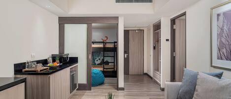 A modern apartment with a kitchen, dining area, and living room. There is a coffee table with a plant, a sofa with cushions, and a blue bean bag. The apartment has wooden flooring and a large window.