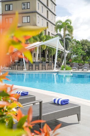 A swimming pool with lounge chairs and a covered seating area, surrounded by palm trees and a multi-story building in the background.