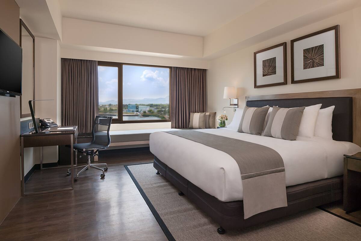 A hotel room with a large bed, a desk with a laptop, a chair, a TV, and a window with a view of buildings and mountains.