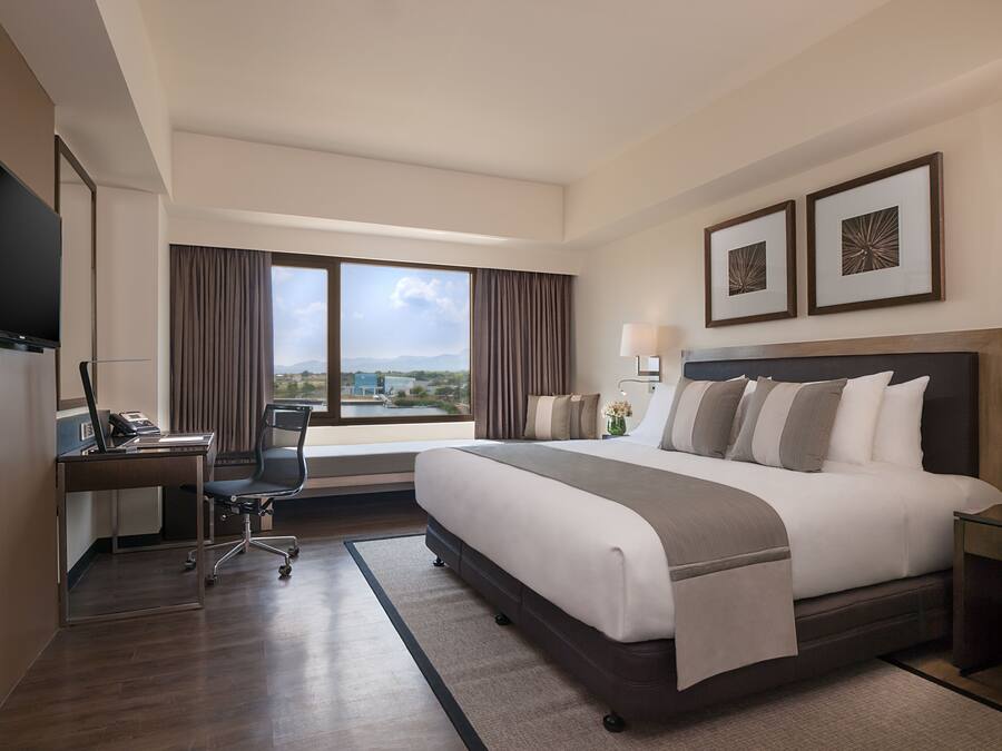 A hotel room with a large bed, a desk with a laptop, a chair, a TV, and a window with a view of buildings and mountains.