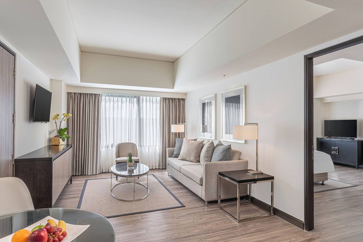 A modern hotel room with a sofa, a small table, a TV, and a dining area with fruit.