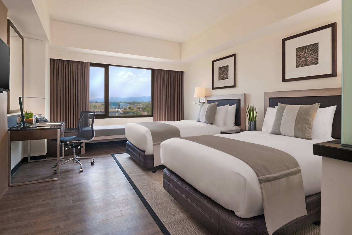 A hotel room with two beds, a desk, a chair, a TV, and large windows with a view.