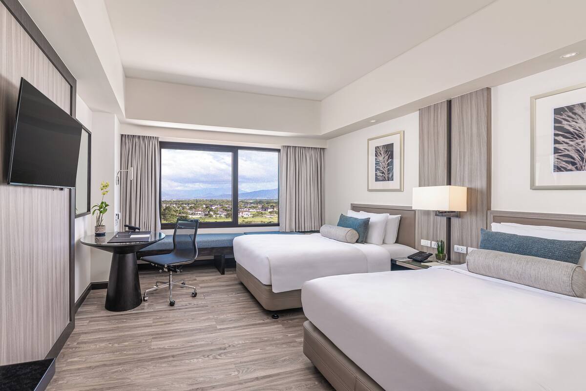 A hotel room with two beds, a desk, a TV, and a large window with a city view.