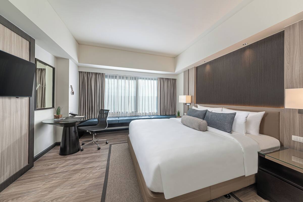A hotel room with a large bed, a desk with a chair, a flat-screen TV, a window with curtains, and a nightstand with a lamp.