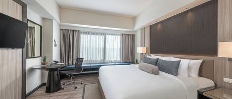 Habitación de hotel con una cama grande, un escritorio con silla, un televisor de pantalla plana, una ventana con cortinas y una mesita de noche con lámpara.