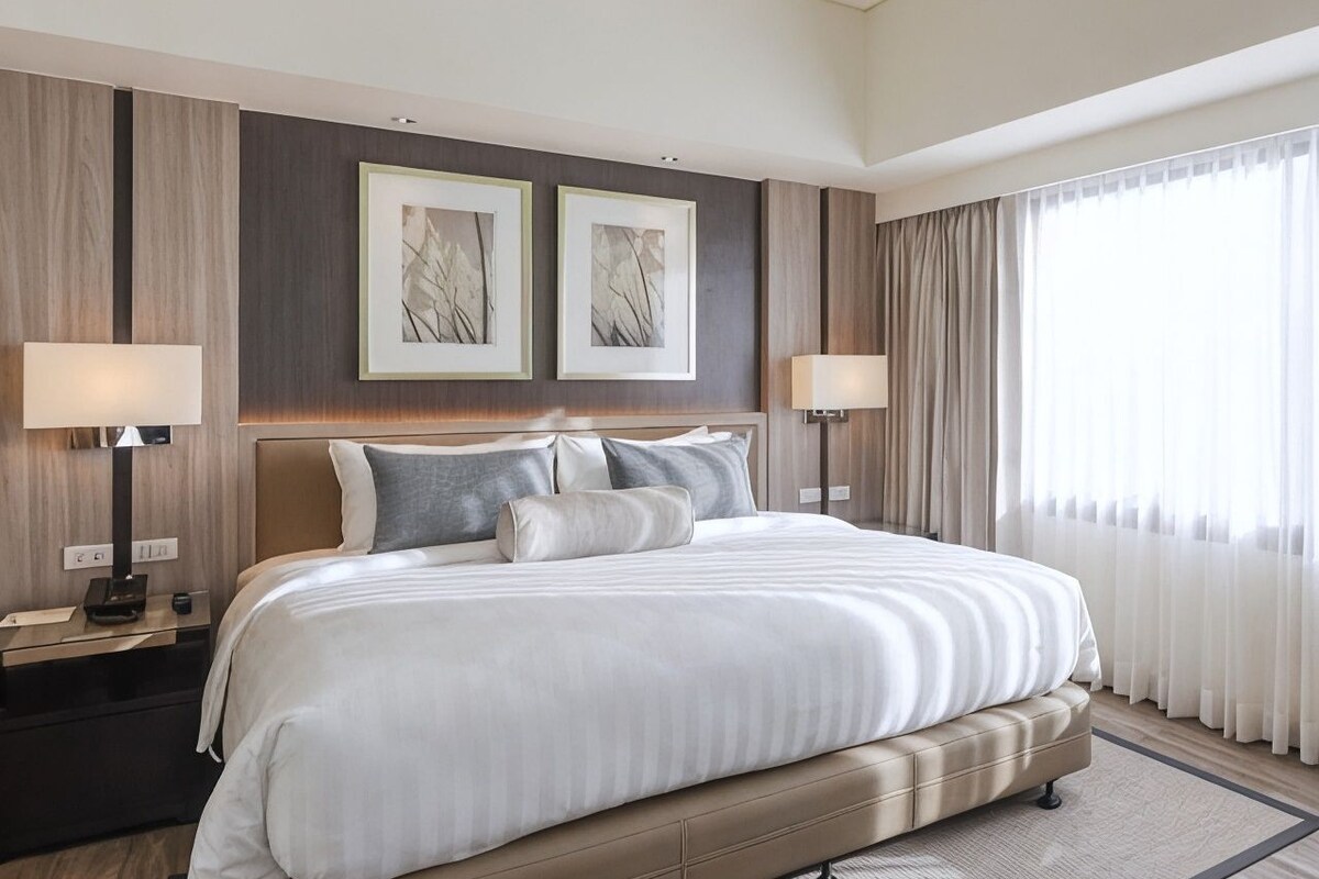 Una habitación de hotel con una cama grande, dos lámparas de noche, un cabecero y una ventana con cortinas.