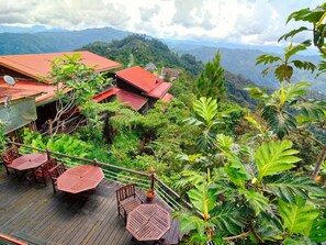 Una terraza de madera con mesas y sillas, rodeada de abundante vegetación y un paisaje montañoso.
