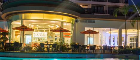 A hotel pool area with outdoor seating and umbrellas, surrounded by a modern building with balconies.