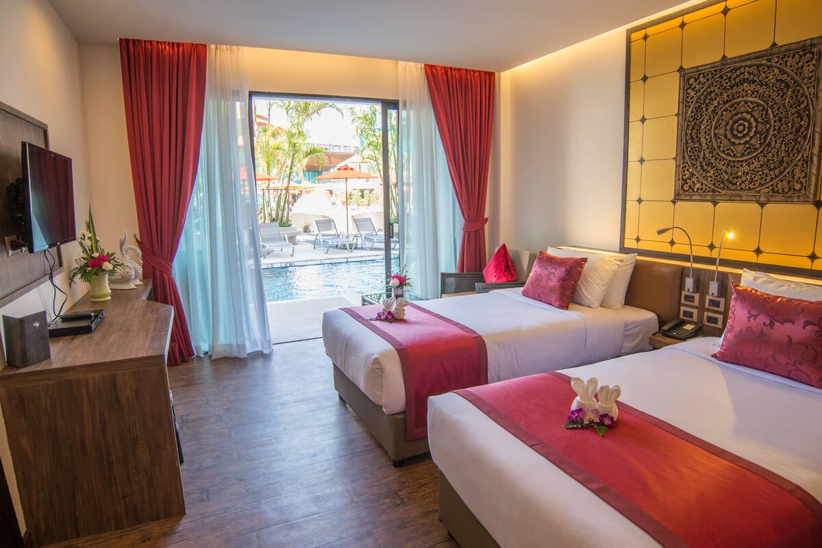 Habitación de hotel con dos camas, un escritorio con televisor, una computadora portátil y vista a una piscina y palmeras.