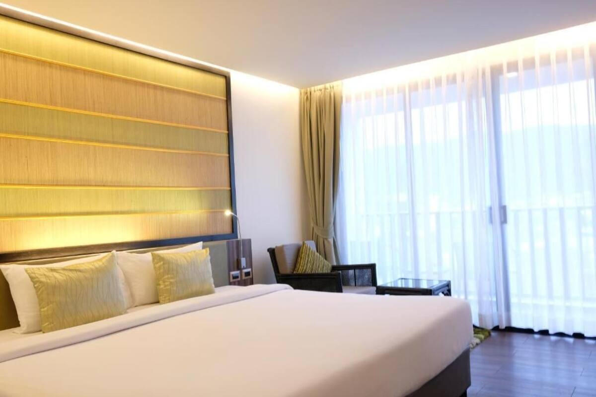 Una habitación de hotel moderna con una cama grande, cabecera con paneles iluminados, zona de estar con una mesa y sillas, y un ventanal con cortinas translúcidas.