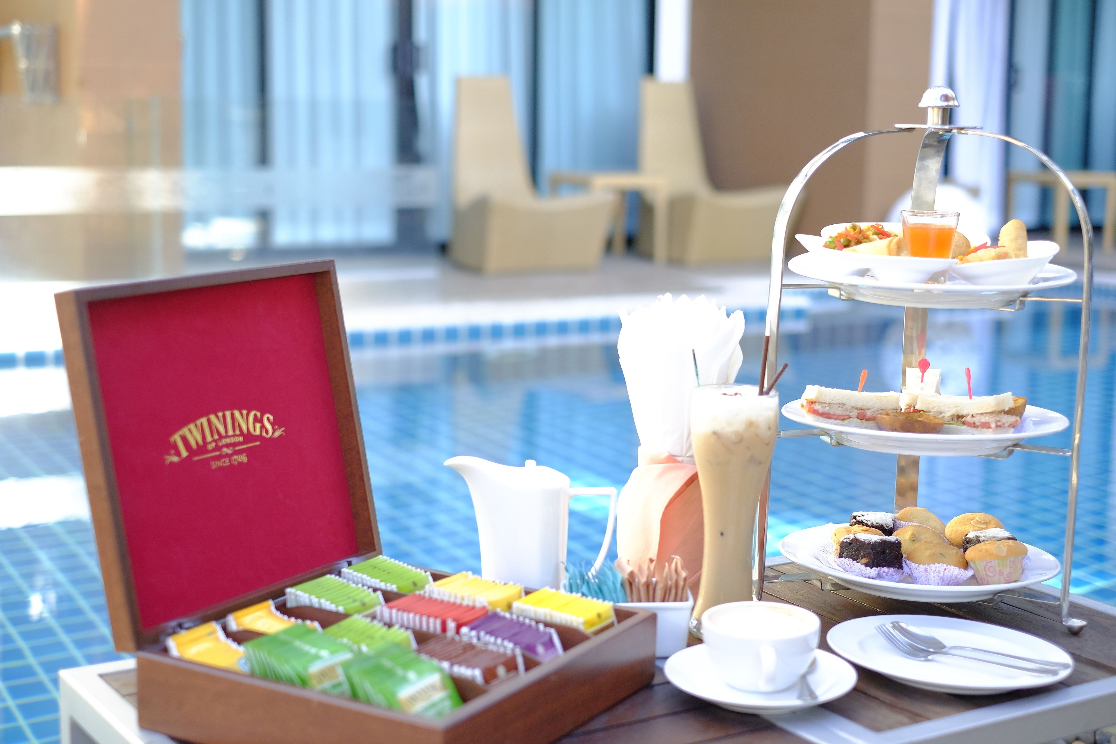 A tray with a tiered dessert stand, a selection of colorful packaged snacks, a pitcher, and a cup, set against a poolside view.
