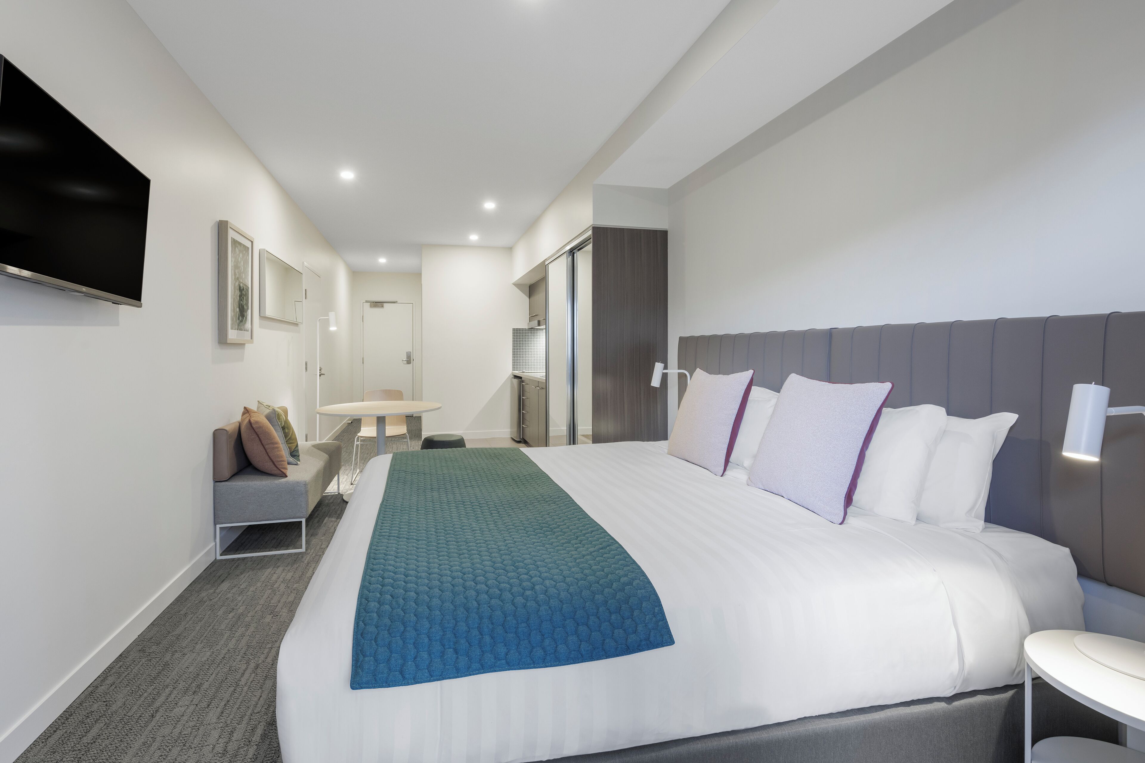 A modern hotel room with a large bed, a flat-screen TV mounted on the wall, a small seating area with a table, and a bathroom visible through an open door.