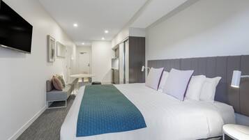 A modern hotel room with a large bed, a flat-screen TV mounted on the wall, a small seating area with a table, and a bathroom visible through an open door.