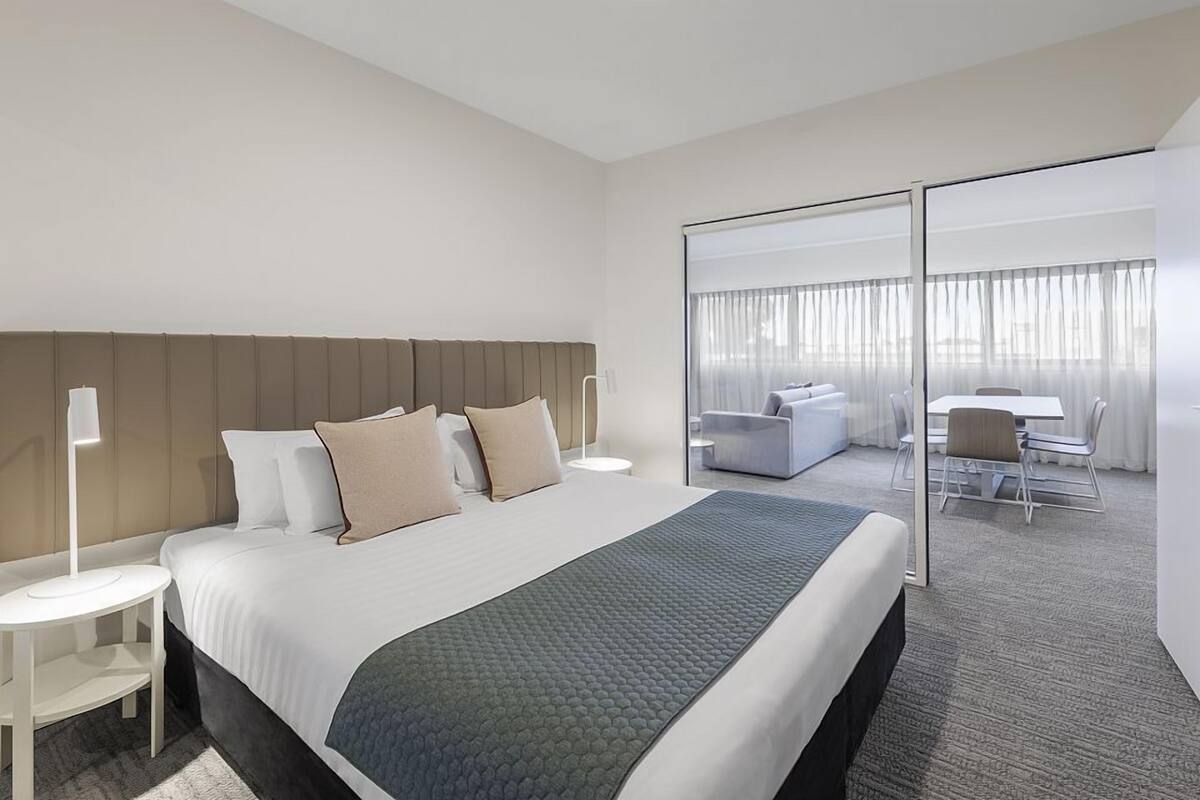 Ein Hotelzimmer mit einem großen Bett, einem Nachttisch, einem Schreibtisch mit Stuhl und Blick durch eine Glastür auf einen Balkon mit einer Couch und einem Tisch.