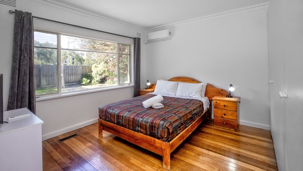 A bedroom with a wooden bed, a nightstand, a window with curtains, and a white wall. - Parkside Inn Motel (Kingsbury)