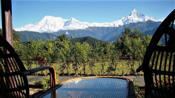 A view from a covered outdoor seating area with a glass table, overlooking a mountainous landscape with snow-capped peaks. - Himalayan Deurali Resort (Pokhara)