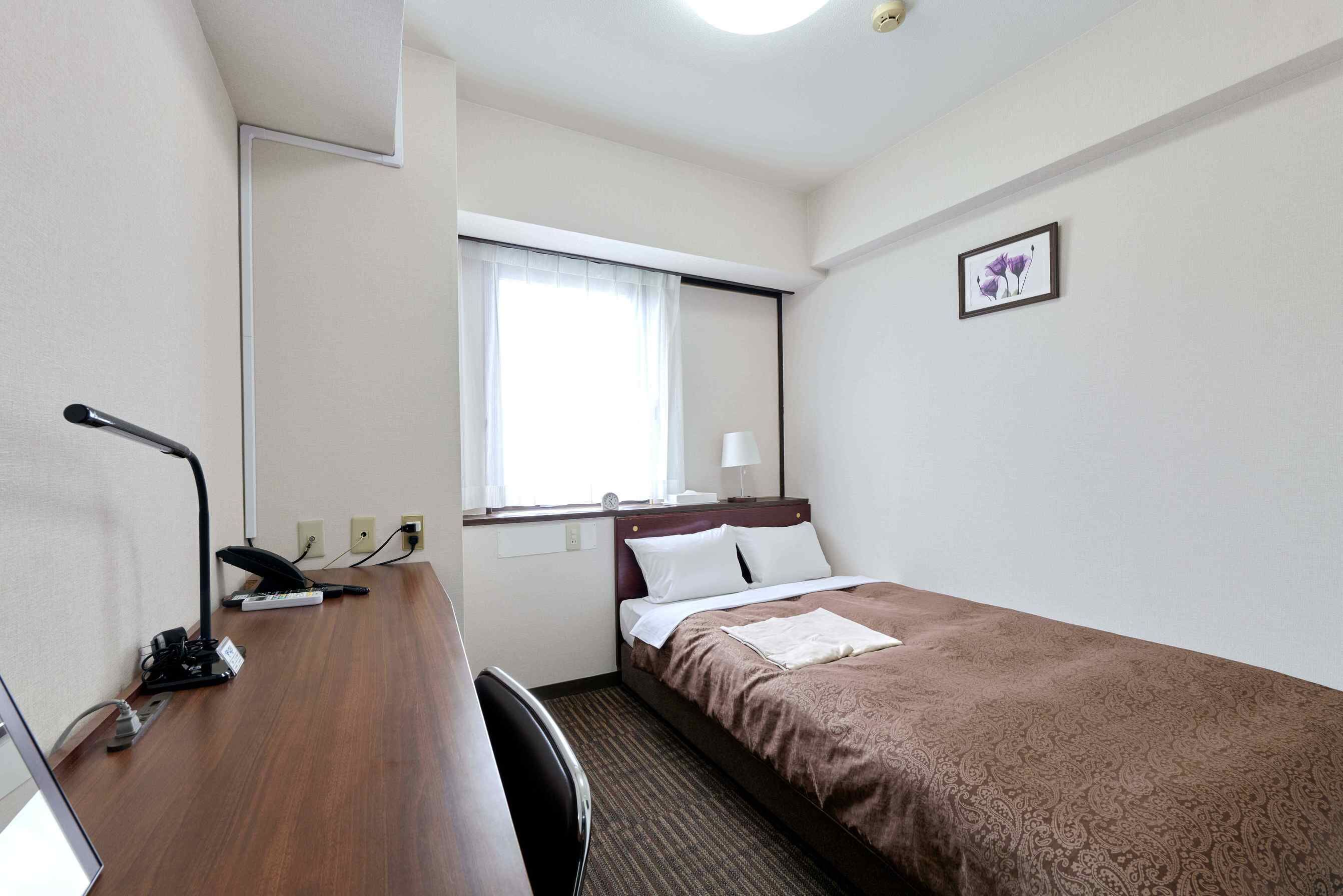 A hotel room with a single bed, a desk with a chair, a window with curtains, a small bedside table, and a wall-mounted picture.