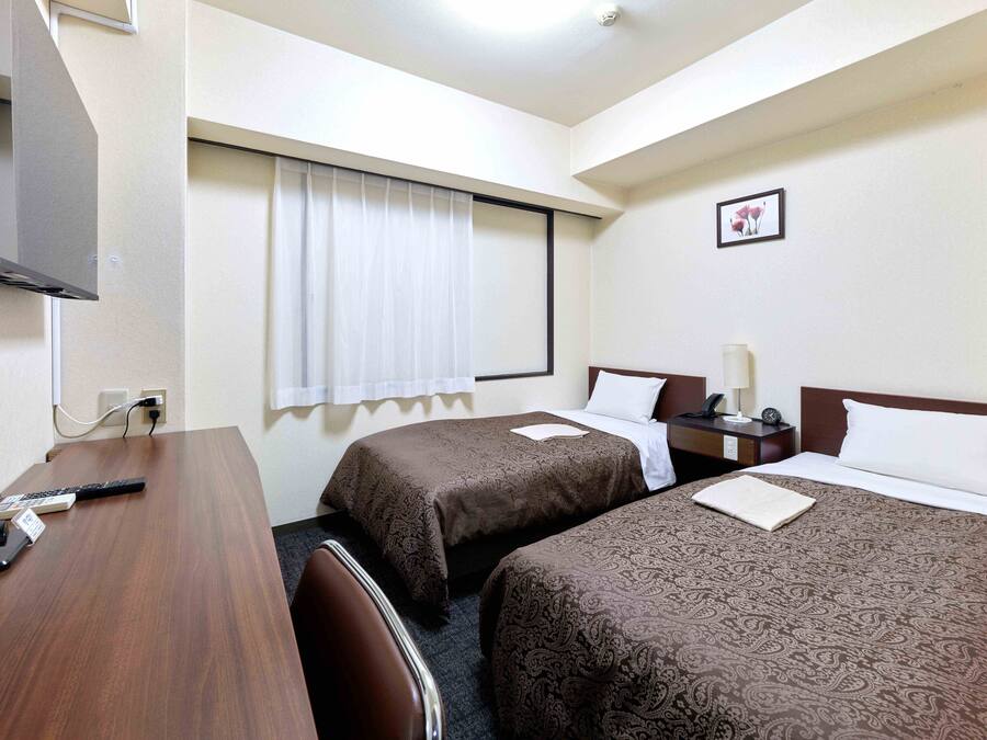 A hotel room with two beds, a desk with a computer and phone, a window with curtains, and a wall-mounted TV.