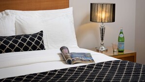 Una cama bien hecha con almohadas blancas y estampadas, una revista, una lámpara y una botella sobre una mesita de noche.